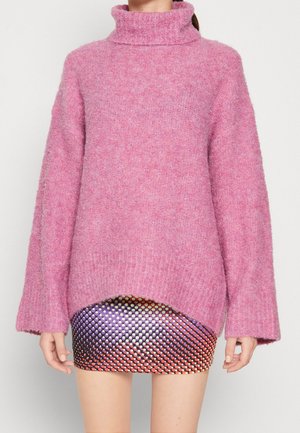 Donna che indossa un maglione dolcevita rosa largo sopra una gonna corta multicolore con motivo a pois, in piedi su uno sfondo bianco.
