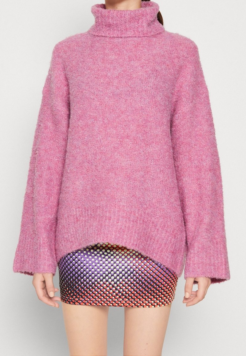 Femme portant un pull à col roulé rose ample sur une jupe courte multicolore à motif à pois, debout sur fond blanc.