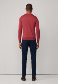 Muž oblečený v přiléhavém červeném dlouhém tričku, tmavě modrých kalhotách a tmavě hnědých botách, stojící čelem k jednobarevnému světle šedému pozadí.