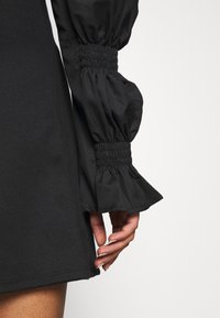 Černé šaty s balónovými rukávy a řasenými manžetami, vyrobené z hladkého materiálu, s fitted korzetem a rozšířeným lemem.