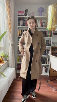 Beige double-breasted coat with large black buttons, side pockets, and a straight silhouette. The fabric appears smooth and tailored.