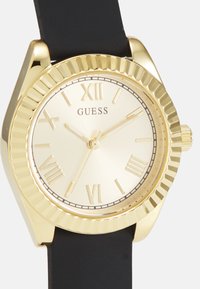 Reloj dorado con esfera redonda, correa de silicona negra, marcadores de números romanos, bisel texturizado y esfera dorada lisa.