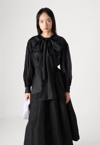 Zwarte blouse met grote strikdetail bij de hals; heeft lange, geplooide mouwen. Gecombineerd met een zwarte, vloeiende rok en een lichtgrijze handtas.