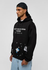 Zwarte hoodie met witte tekst, voorzien van een voorzak met blauwe en witte verfspatten. Zachte stof, met trekkoord in de capuchon, comfortabele pasvorm.