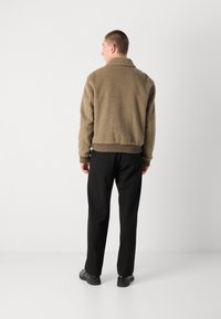 Braune strukturierte Jacke mit Kragen, geripptem Saum und schwarzen Hosen. Das Outfit umfasst schwarze Schuhe und wird von hinten betrachtet.