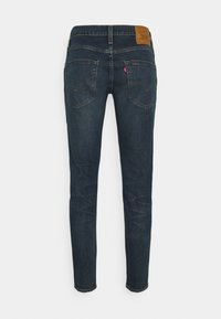 Jean skinny bleu foncé vu de dos avec deux poches arrière, un patch en cuir de la marque sur la ceinture et une petite étiquette rouge sur la poche droite.