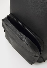 Zaino in pelle nera con una superficie testurizzata, una tasca frontale con zip e un pannello liscio per il branding nella parte inferiore.