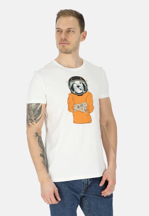 Mand med tatoveret arm iført hvid T-shirt med grafik af en Dalmatinerhund i en orange rumdragt, kombineret med blå jeans.