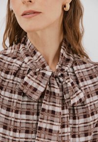 Braun-und-beige-karierte Bluse mit gebundenem Kragen. Verfügt über eine weiche Textur und leichten Stoff, ergänzt durch einen subtilen Glanz.