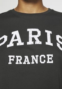 Camiseta de algodón gris con cuello redondo, que presenta un texto en blanco audaz y desgastado que dice "PARÍS FRANCIA". Textura suave y diseño casual.