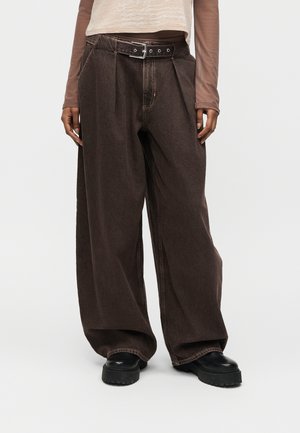 Osoba ubrana w luźne, ciemnobrązowe spodnie o szerokich nogawkach z paskiem, czarne masywne buty i beżowy długi rękaw z przezroczystymi rękawami.