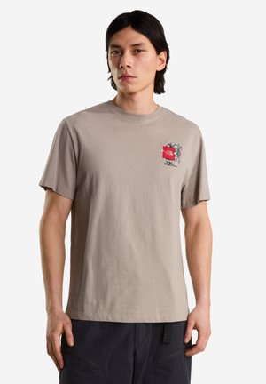 Joven con una camiseta beige de manga corta con un pequeño gráfico rojo y negro en el pecho izquierdo, combinada con pantalones oscuros.
