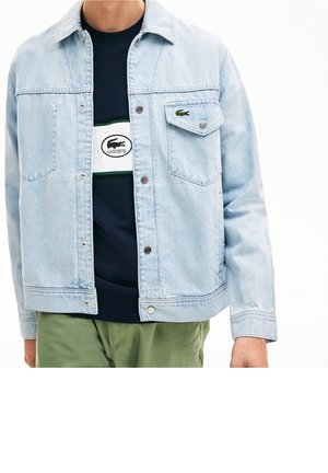 Osoba nosząca jasnoniebieską kurtkę dżinsową na granatowej bluzie Lacoste z logo, w połączeniu z jasnzielonymi spodniami.