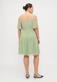 Lichtgroene jurk met een getailleerde-bovenkant, zwierige rok, doorschijnende mouwen met koude schouders en een V-rug. Gecombineerd met zwarte slingback hakken.