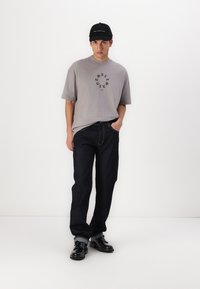 Camiseta gris de gran tamaño con gráfico circular negro, jeans rectos de mezclilla azul oscuro y gorra negra. Zapatos negros con detalles de hardware en plata.