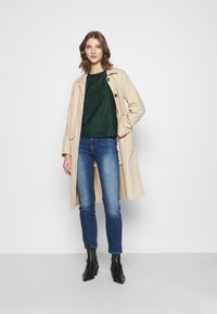 Casaco trench bege com um design estruturado, top de renda verde, jeans azuis e botas de tornozelo pretas com salto, contra um fundo branco liso.