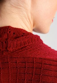 Strickpullover in tiefrot mit einem strukturierten Muster. Verfügt über einen gerippten Halsausschnitt mit detaillierten Nähten und offenen Webbereichen für Atmungsaktivität.