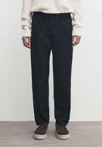 Marinblå elastiska midjebyxor i rak modell, tillverkade av slät tyg. I kombination med bruna mockaskor med vita sulor.