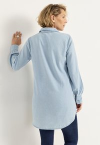 Ljusblå denimskjorta med krage, långa ärmar, knappmanschetter och en rundad nederkant, överdragen bakifrån mot en vit bakgrund.