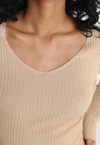 Béžový žebrovaný top s širokým V-výstřihem, dlouhými rukávy a texturovanou tkaninou, která zdůrazňuje jeho přiléhavý tvar.