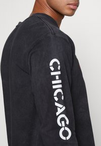 Svart långärmad bomullströja med vit text "CHICAGO" på ärmen och en subtil sliten textur. Rund halsringning och avslappnad passform.