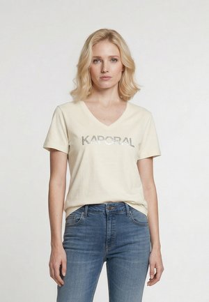 Blondynka stoi przodem, ubrana w kremowy T-shirt z dekoltem w serek z napisem „KAPORAL” oraz niebieskie spodnie jeansowe z wysokim stanem na jednolitym białym tle.
