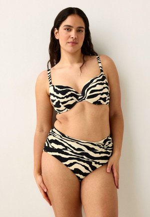 Kvinde med langt mørkt hår iført sort-hvid zebra-print bikini, stående mod en ensartet lys baggrund.