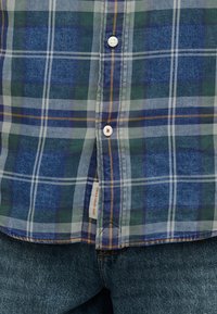 Καρό πουκάμισο σε αποχρώσεις μπλε, πράσινου και λευκού, κουμπωμένο πάνω από σκούρα μπλε τζιν, με μια μπεζ ετικέτα που έχει πορτοκαλί κείμενο κοντά στο κάτω μέρος του πουκαμίσου.