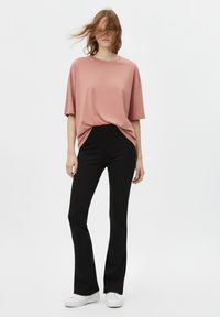 Maglietta rosa oversize con maniche corte infilata in pantaloni neri a zampa ad alta vita, abbinata a sneakers bianche. Tessuto dalla texture liscia.