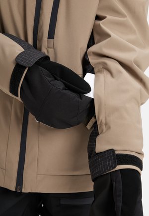 Person som bär en beige jacka och svarta handskar lägger ett vitt kort i en dragkedja på jackans ärm.