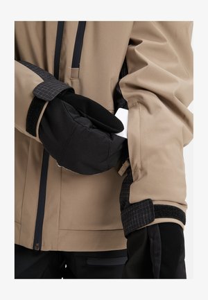 Osoba v béžové bundě a černých rukavicích vkládá bílou kartu do zipové kapsy na rukávu bundy.