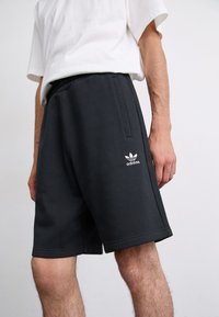 Black cotton shorts with an elastic waistband and side pockets. Features a white Adidas logo on the left leg. Short hem, relaxed fit.