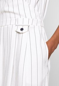 Main dans la poche d’un vêtement blanc à fines rayures verticales noires et un passant de ceinture avec un bouton noir.
