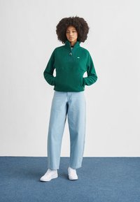 Grønn fleecegenser med kvartglidelås og frontlomme, kombinert med lyseblå høytlivsjeans og hvite joggesko.