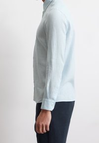 Uomo in camicia azzurra a maniche lunghe e pantaloni scuri in piedi di lato contro uno sfondo bianco uniforme.