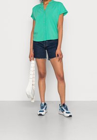 Blusa corta de manga corta color turquesa con escote en V, combinada con pantalones cortos de mezclilla. Bolso blanco con tachuelas y zapatillas azules con detalles de malla.