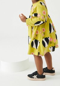 Grünes Kleid mit schwarzem und weißem Panda-Muster, lange Ärmel, ausgestellter Rock. Schwarze Glitzer-Sneaker mit einem pinken Stern-Akzent.