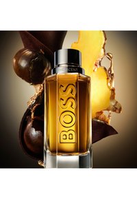 Bottiglia di profumo in vetro con forma rettangolare, liquido di colore ambra, etichetta in rilievo con la scritta "BOSS" e un tappo d'argento, circondata da zenzero e baccelli di cacao.