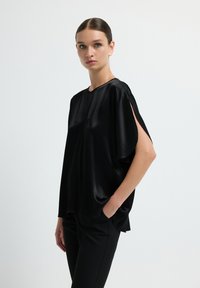 Top in satin nero con vestibilità ampia, maniche corte e delicate fessure laterali. Abbinato a pantaloni neri eleganti per un look minimalista.