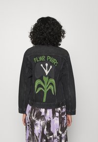 Person med lockigt hår bär en svart denimjacka med grön och vit blommönstring och texten "FLWR PWRS" på ryggen, ihop med en lila mönstrad kjol.