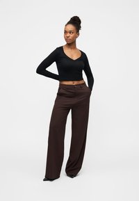 Νεαρή γυναίκα που στέκεται με το ένα χέρι πίσω από την πλάτη και το άλλο στην τσέπη, φορώντας μαύρο μακρυμάνικο κοντό τοπ και καφέ παντελόνι με φαρδιά μπατζάκια.