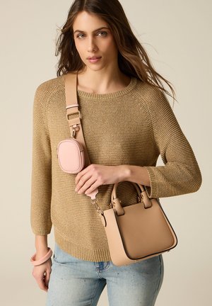 Nainen, jolla on beige neulepusero ja vaaleansiniset farkut, pitää kädessään vaaleanruskeaa käsilaukkua, jossa on leveä hihna ja pieni vaaleanpunainen pussi kiinnitettynä rinnan yli.