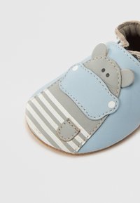 Zapatito suave para bebé en cuero azul claro, con un diseño de cara de animal gris, patrón a rayas y punta redondeada. Incluye costuras decorativas.
