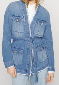 Light blue denim jacket with a tie waist, two front pockets, snap button accents, and frayed hem details, layered over a white collared shirt.