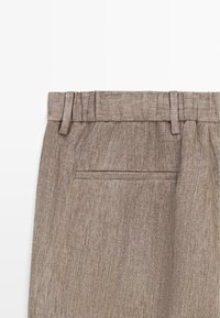 Primo piano della vita di pantaloni di lino beige con passanti per cintura e una tasca a filetto singola, su sfondo bianco.