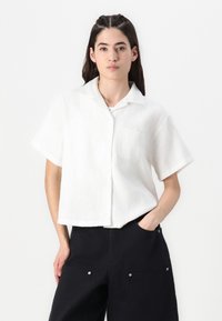 White short-sleeve button-up shirt with a textured pattern, featuring a chest pocket and a relaxed fit. Worn with black pants.