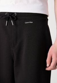 Černé tepláky vyrobené z bavlněné směsi, s pasem na šňůrku, bočními kapsami a malým logem "Calvin Klein" na bocích.
