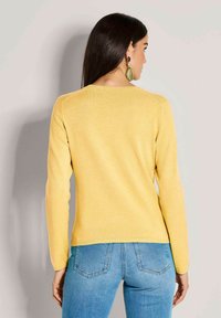 Gelber Strickpulli mit langen Ärmeln, Rundhalsausschnitt und geripptem Saum. Kombiniert mit blauen Jeans, die eine enganliegende Silhouette zeigen.