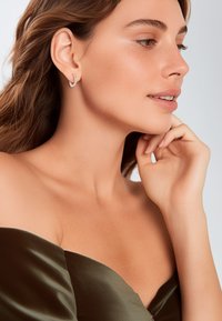 Zilveren oorringen met kleine gekleurde edelstenen, die een gebogen ontwerp laten zien. Model draagt een donkere satijnen off-shoulder top. Glad huidtextuur.