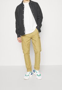 Neformální oblečení s černou košilí na knoflíky, bílým tričkem, khaki cargo kalhotami a bílými teniskami s barevnými akcenty.